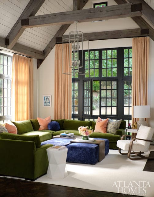Living room with vaulted wood-beamed ceiling, large black-paned window overlooking greenery, and a large sectional sofa in olive green with blue and coral accents.