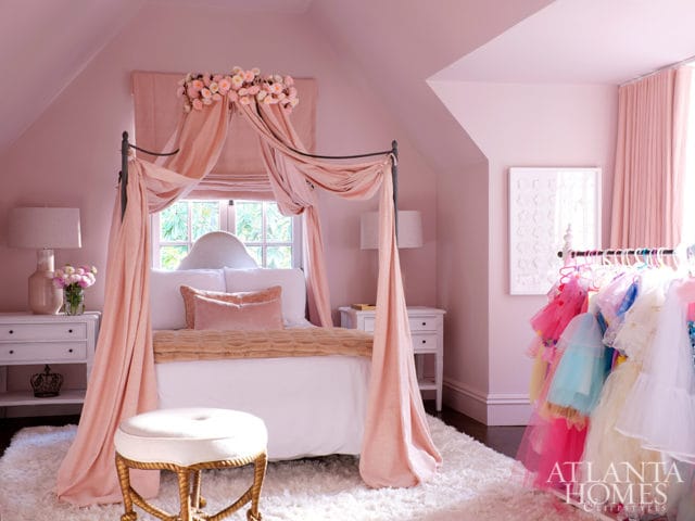 Pink bedroom with a canopy bed draped in sheer pink fabric, a white shag rug, and a rack of colorful children's dresses.
