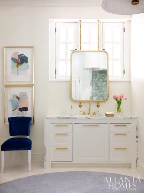 White bathroom vanity with marble countertop and gold hardware, flanked by two framed abstract art pieces and a navy velvet chair.