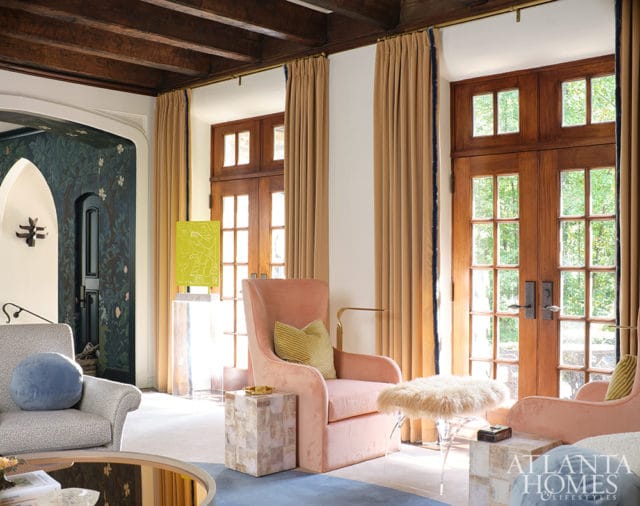 Living room with plush pink armchairs, a patterned armchair, and wooden French doors leading outside. Dark floral wallpaper adorns an arched doorway.