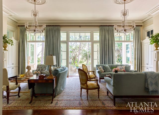 Light blue sofas and chairs arranged in a living room with tall windows and doors opening to an outdoor patio. Crystal chandeliers hang from the ceiling.