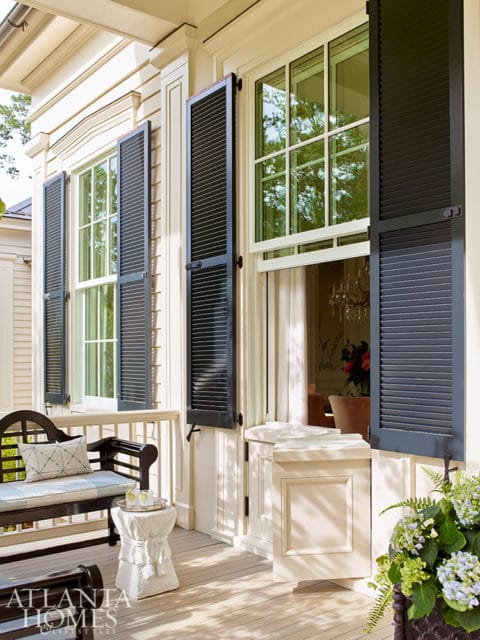 Cream-colored house exterior with black louvered shutters on multiple windows, a dark wood bench with cushions, and a white garden stool on a wooden porch.