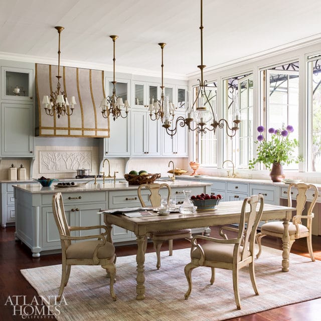 Light blue kitchen with a large island, marble countertops, brass fixtures, and a distressed wooden dining table with upholstered chairs.