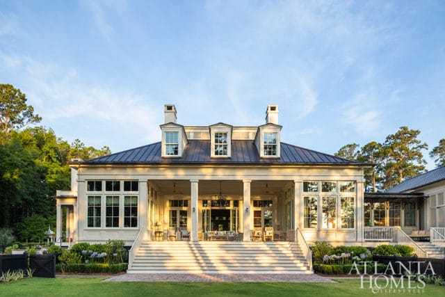 Large cream-colored house with a dark metal roof, multiple dormer windows, and a wide covered porch with seating. A grand staircase leads up to the porch, with manicured gardens and trees surrounding the property.
