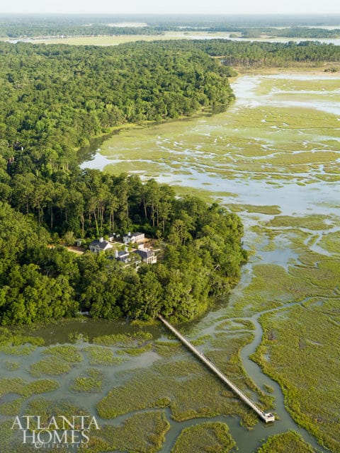 Aerial view of a white house with multiple buildings nestled in a lush green forest, overlooking a marshy waterway with a long pier extending into the water.