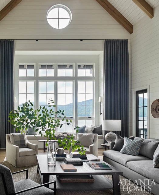 Vaulted ceiling living room with shiplap walls, exposed beams, and a large window overlooking a lake. Furnishings include a gray sofa, armchairs, and a dark wood coffee table.