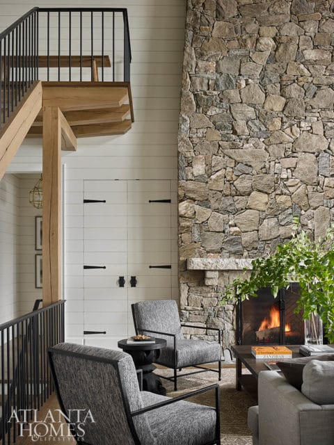 Living room with a large stone fireplace, two gray upholstered armchairs, and a wooden staircase with black railings.