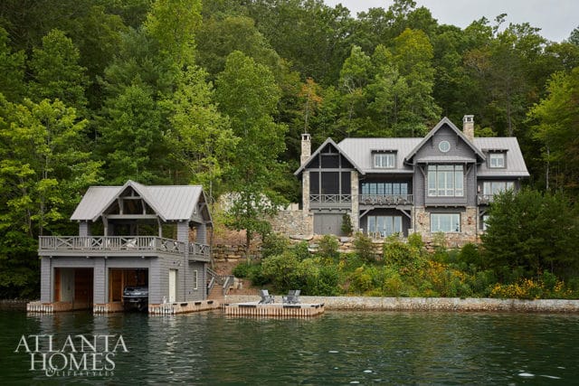 Gray lake house with metal roof and stone accents, featuring a boathouse with garage and a wooden dock with chairs. Surrounded by lush green trees.