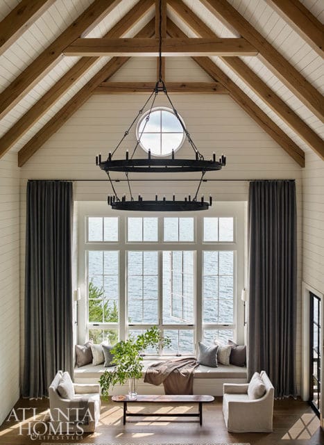 Vaulted ceiling living room with exposed wooden beams, shiplap walls, and a large window overlooking the water. Features a two-tiered black chandelier, gray curtains, and a built-in window seat with plush cushions.