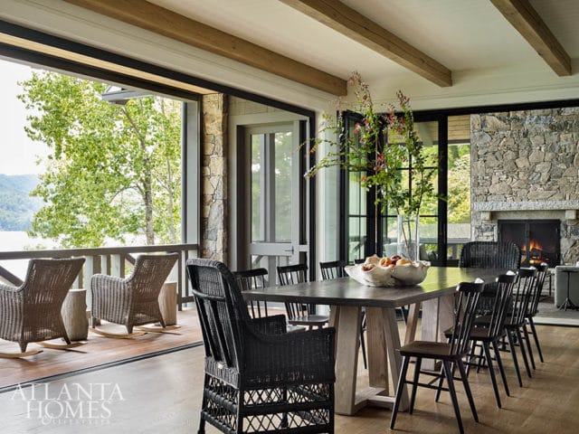 Dining room with a large stone fireplace, wooden beams, and a table set for a meal, overlooking a lake and trees through open doors.
