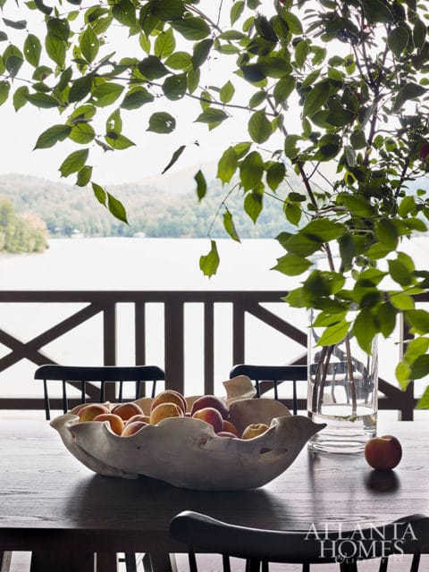 Wooden dining table with a large, organic-shaped bowl filled with peaches, a glass vase with greenery, and a single peach. Chairs are visible in the foreground and background, with a lake and mountains seen through a wooden railing.