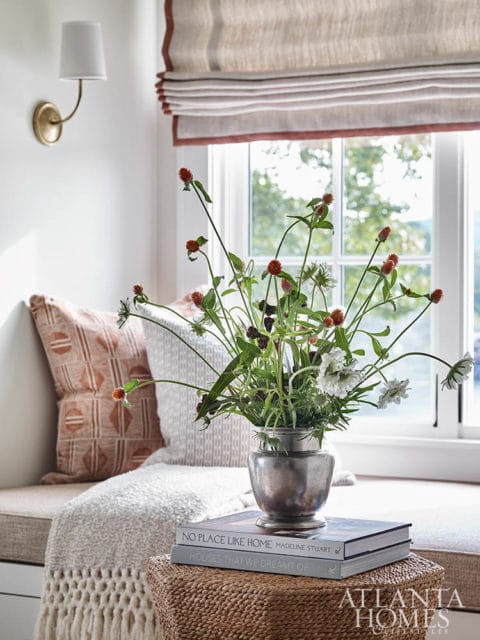 Window seat with neutral cushions and pillows, a metal vase of wildflowers, and a stack of design books on a woven stool.
