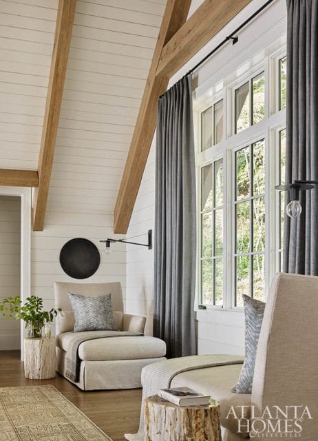 Living room with vaulted shiplap ceiling, exposed wood beams, and large windows. Features two upholstered chairs, gray curtains, and rustic tree stump side tables.