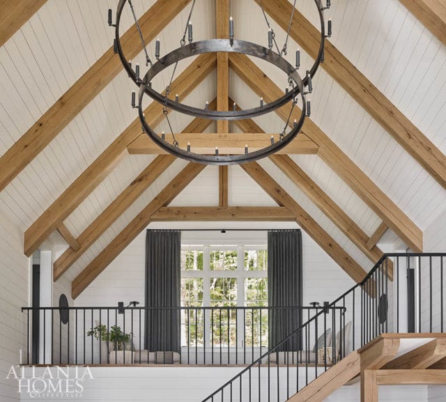 Vaulted ceiling with exposed wooden beams and white shiplap walls, featuring a large circular chandelier and a second-floor balcony with a black railing overlooking a window with dark gray curtains.