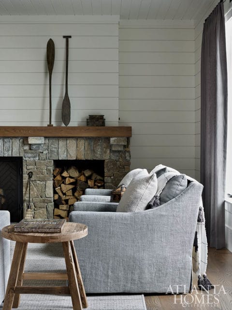 Living room with stone fireplace, shiplap walls, and a gray upholstered sofa with decorative pillows and a throw blanket. Two wooden oars hang above the mantel.