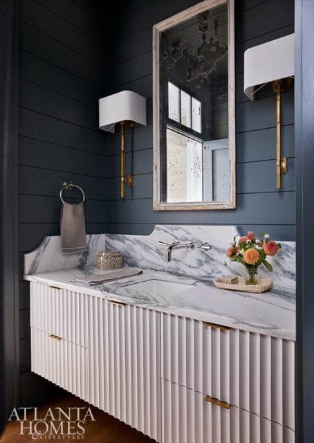 Powder room with dark blue shiplap walls, marble vanity with fluted white cabinetry, brass sconces, and a distressed mirror.