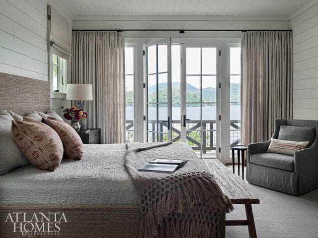 Bedroom with shiplap walls, a king-sized bed with textured headboard, and double doors opening to a lake view. Gray armchair and wooden bench are visible.