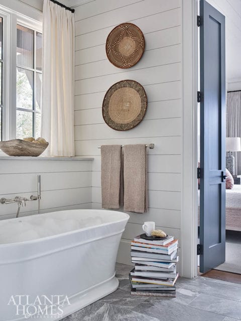 White freestanding bathtub with chrome faucet, shiplap walls adorned with two woven baskets, and a stack of magazines next to a blue door opening to a bedroom.