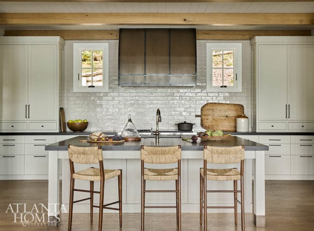 White kitchen with a large gray island, three woven bar stools, white subway tile backsplash, and a stainless steel range hood.