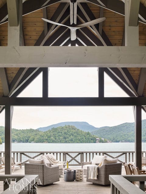 Covered porch with exposed wooden beams and a ceiling fan, overlooking a lake and tree-covered mountains.