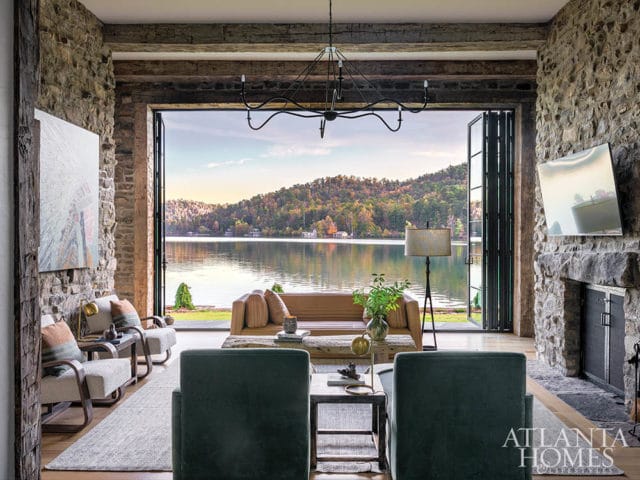 Living room with stone walls, large folding glass doors opening to a lake view, leather sofa, armchairs, and a stone fireplace.