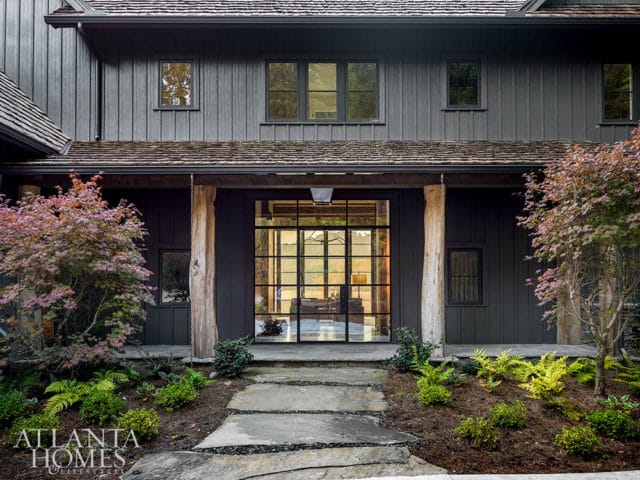 Dark wood-sided house with a large glass-paned front door, natural wood support beams, and lush landscaping with ferns and a Japanese maple tree.