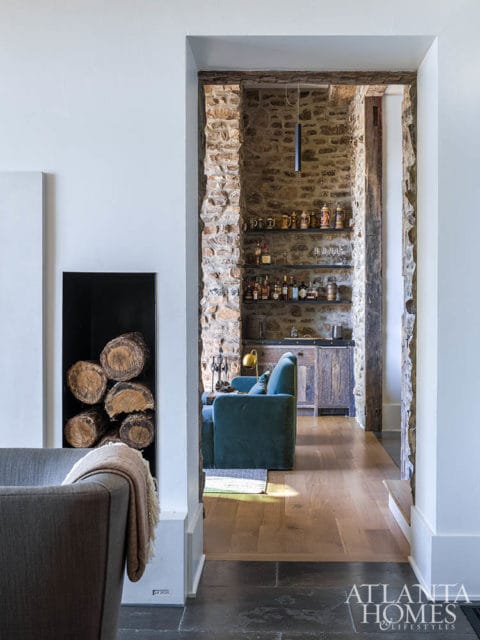 View through an archway into a bar area with stone walls, shelves stocked with bottles and mugs, and a teal sofa. A wood-paneled cabinet sits below the shelves.