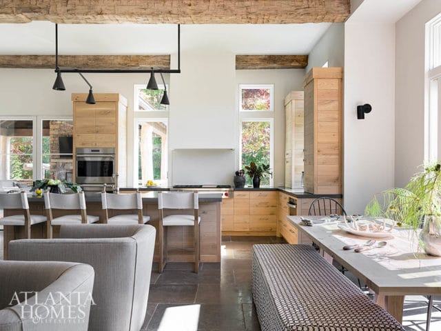 Open-plan kitchen with natural wood cabinetry, stone floors, exposed wooden beams, and a dining area with a light wood table and patterned bench.
