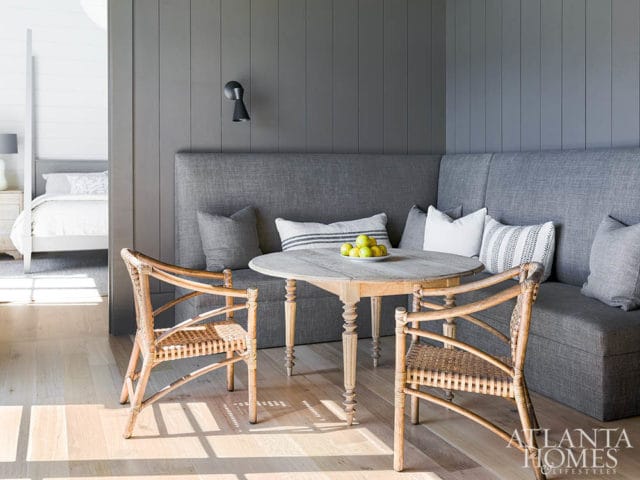 Gray upholstered banquette seating with patterned pillows around a round wooden table with two wicker chairs. A bedroom is visible through an opening to the left.