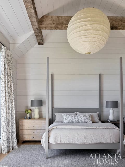 Bedroom with a grey four-poster bed, white shiplap walls, exposed wooden beams, and a large paper lantern light fixture.