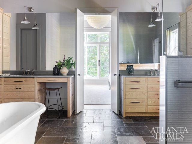 Modern bathroom with dual vanities featuring light wood cabinetry, stone countertops, and brushed metal fixtures. A freestanding tub is visible in the foreground, with a doorway leading to a sunlit room beyond.