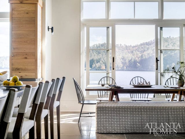 Dining area with a long wooden table, upholstered bench, and chairs, featuring a large window overlooking a lake and wooded hills. A wooden cabinet is visible on the left.