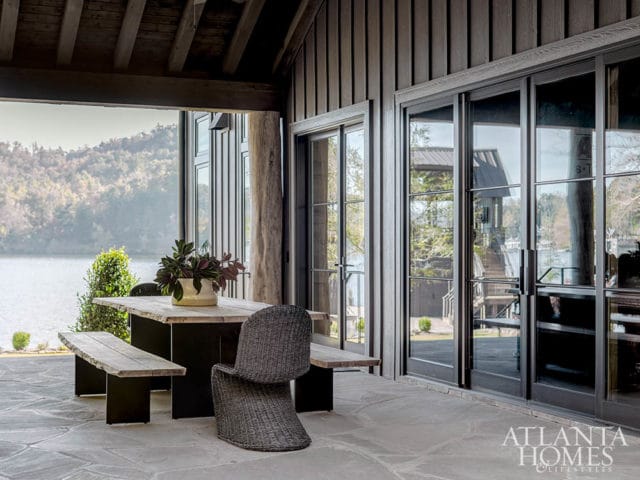 Covered patio with a rustic wooden dining table and benches, wicker chairs, and large sliding glass doors overlooking a lake and wooded hills.