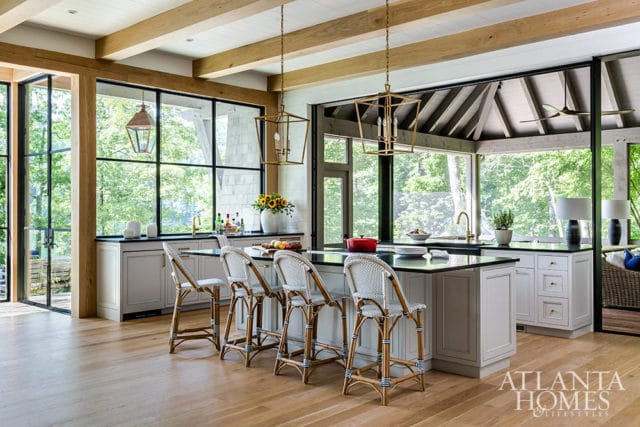 Kitchen with light wood floors, gray cabinets, black countertops, and a large island with white wicker bar stools. Large windows overlook a wooded area.