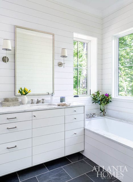 White shiplap bathroom with a double vanity, marble countertops, a soaking tub, and large windows overlooking trees.