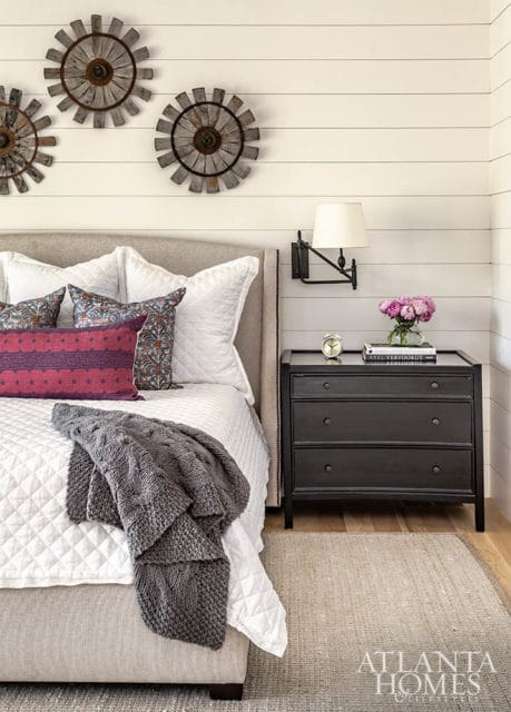Bedroom with shiplap walls, upholstered headboard, quilted white bedding, decorative pillows, gray knit throw, black nightstand with lamp and flowers, and rustic wall decor.