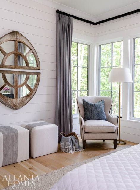 Corner of a bedroom with white shiplap walls, a wingback chair with a blue pillow, two striped ottomans, and a large arched mirror.