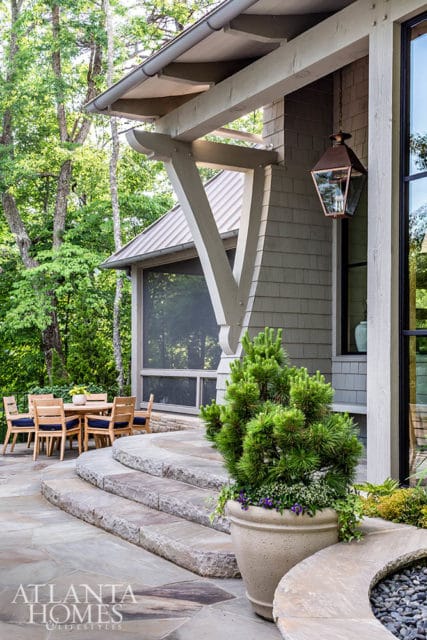 Stone patio with wooden dining set and screened porch, surrounded by trees. Large stone steps lead up to the house with a copper lantern hanging from the porch.