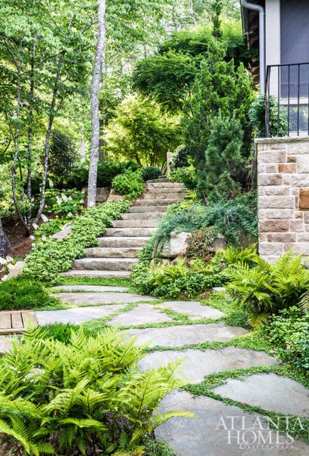 Stone pathway and steps winding through lush greenery and ferns, leading up to a stone-clad house with a balcony.