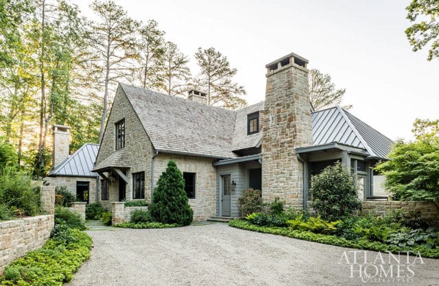 Stone and wood-shingled house with multiple gabled roofs, stone chimneys, and a metal roof section, surrounded by lush greenery and a gravel driveway.