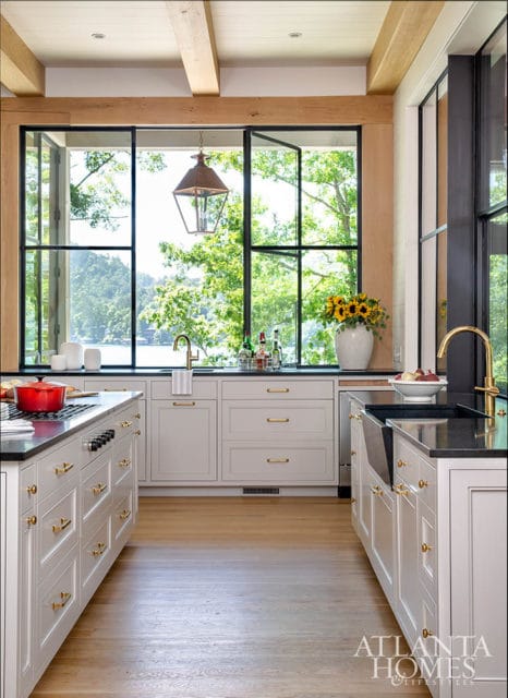 Light gray kitchen cabinets with brass hardware and black granite countertops, featuring a large window overlooking a lake and trees.