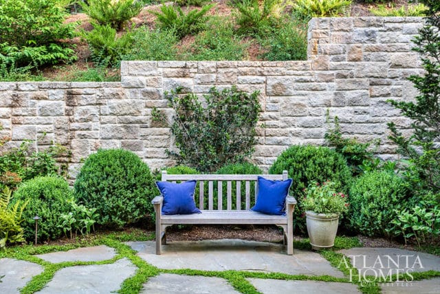 Stone retaining wall with lush green landscaping, a wooden bench with blue cushions, and flagstone patio with grass inlays.