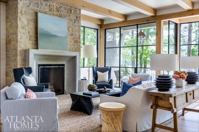 Living room with stone fireplace, blue armchairs, striped sofa, and large windows overlooking trees. Atlanta Homes & Lifestyles logo in bottom left.