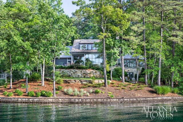 Modern lake house with stone retaining walls and large windows, surrounded by mature trees and lush landscaping, viewed from the water.