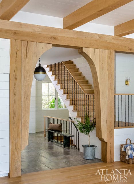 Interior hallway with exposed wooden beams, a wooden staircase with black railings, a dark green bench with a brown cushion, and a potted plant.