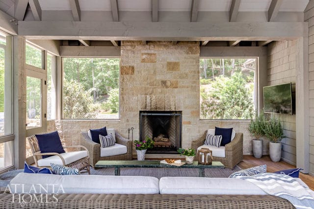 Screened porch with stone fireplace, wicker seating, and a large sectional sofa. Large windows overlook a wooded area.