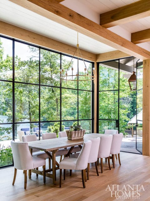 Dining room with a large wooden table and upholstered chairs, overlooking a lake through floor-to-ceiling windows with exposed wooden beams overhead.