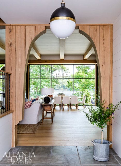 Open-concept living and dining area with large windows overlooking a lake, featuring a neutral-toned sofa, rustic wooden beams, and a modern pendant light.