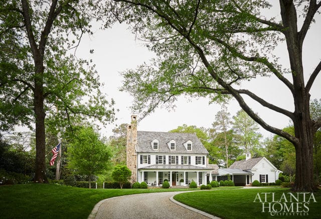 White farmhouse with stone chimney, wraparound porch, and dormer windows, set on a manicured lawn with mature trees and a gravel driveway. Atlanta Homes & Lifestyles logo in the lower right.