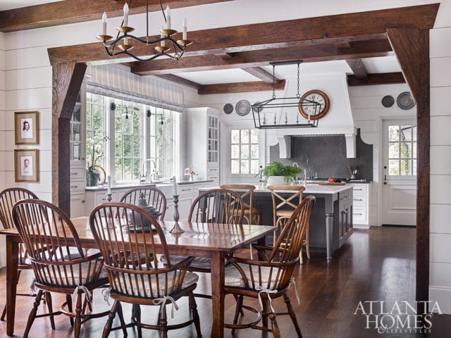 Farmhouse kitchen and dining area with wood beams, shiplap walls, a large wooden dining table with Windsor chairs, and a gray island with a white countertop.